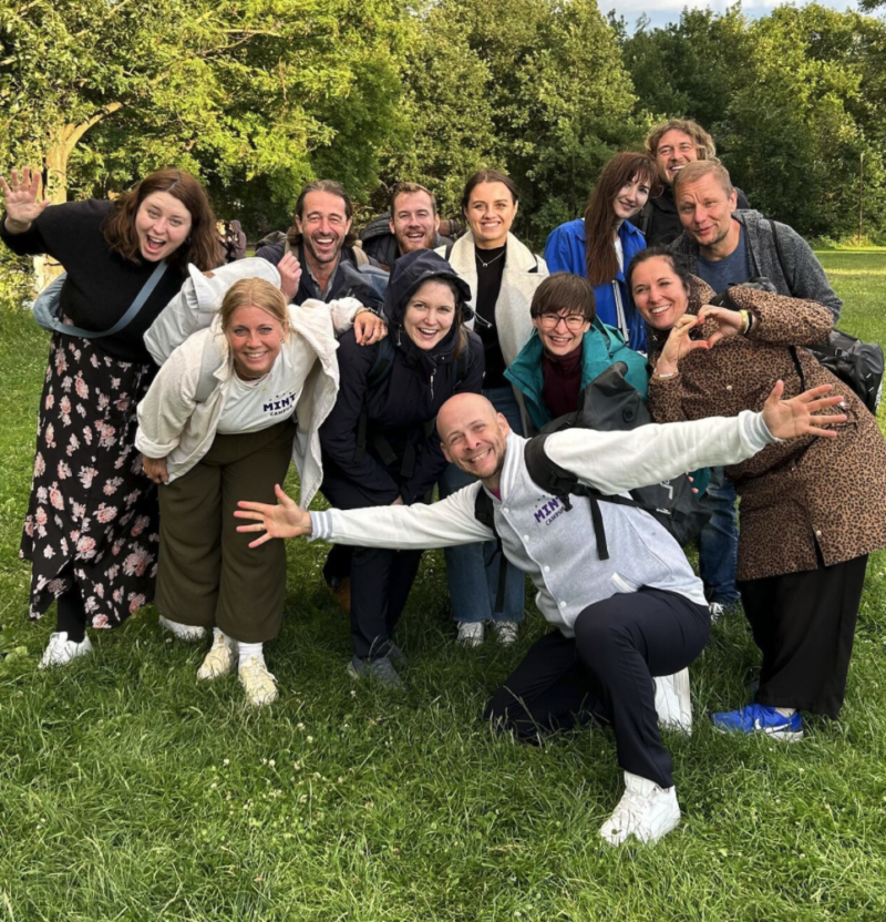 Eine Gruppe von dreizehn Personen sitzt im Freien im Gras und lächelt in die Kamera. Bäume und Grünpflanzen bilden den Hintergrund, und alle sind leger in Jacken und Schichten gekleidet. Werde Teil des Teams und erlebe diesen Teamgeist selbst!.