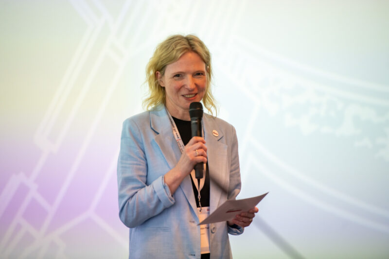 Eine Frau mit blondem Haar, die einen hellblauen Blazer und ein schwarzes Hemd trägt, hält ein Mikrofon und ein Blatt Papier, während sie auf einer Veranstaltung spricht. Der sanft beleuchtete Hintergrund mit abstrakten Mustern unterstreicht ihre Botschaft: Werde Teil des Teams.