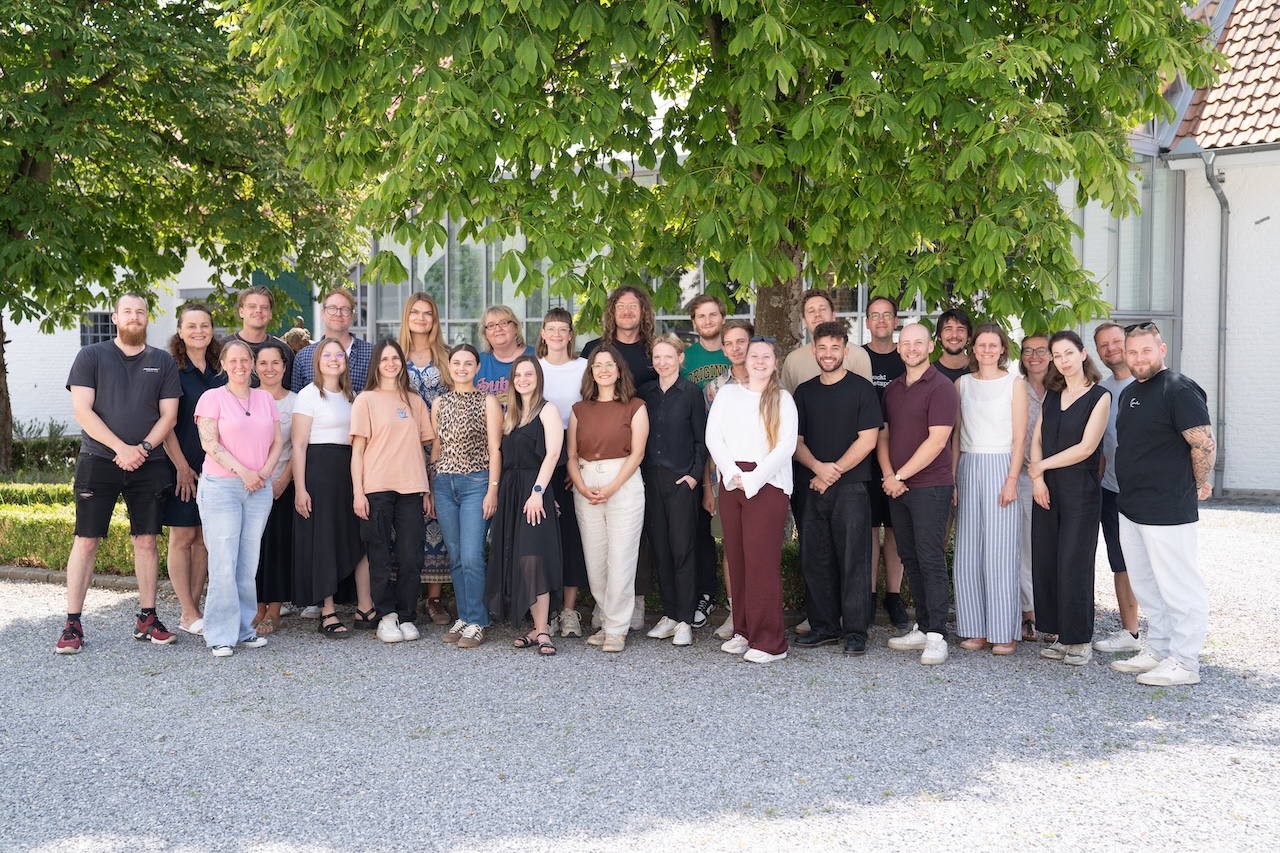 Eine Gruppe von etwa 30 Personen steht an einem sonnigen Tag im Freien zusammen und posiert für ein Foto unter einem großen Baum. Sie lächeln in legerer Sommerkleidung, hinter ihnen ein weißes Gebäude. Werde Teil des Teams und erlebe diesen Zusammenhalt selbst!.